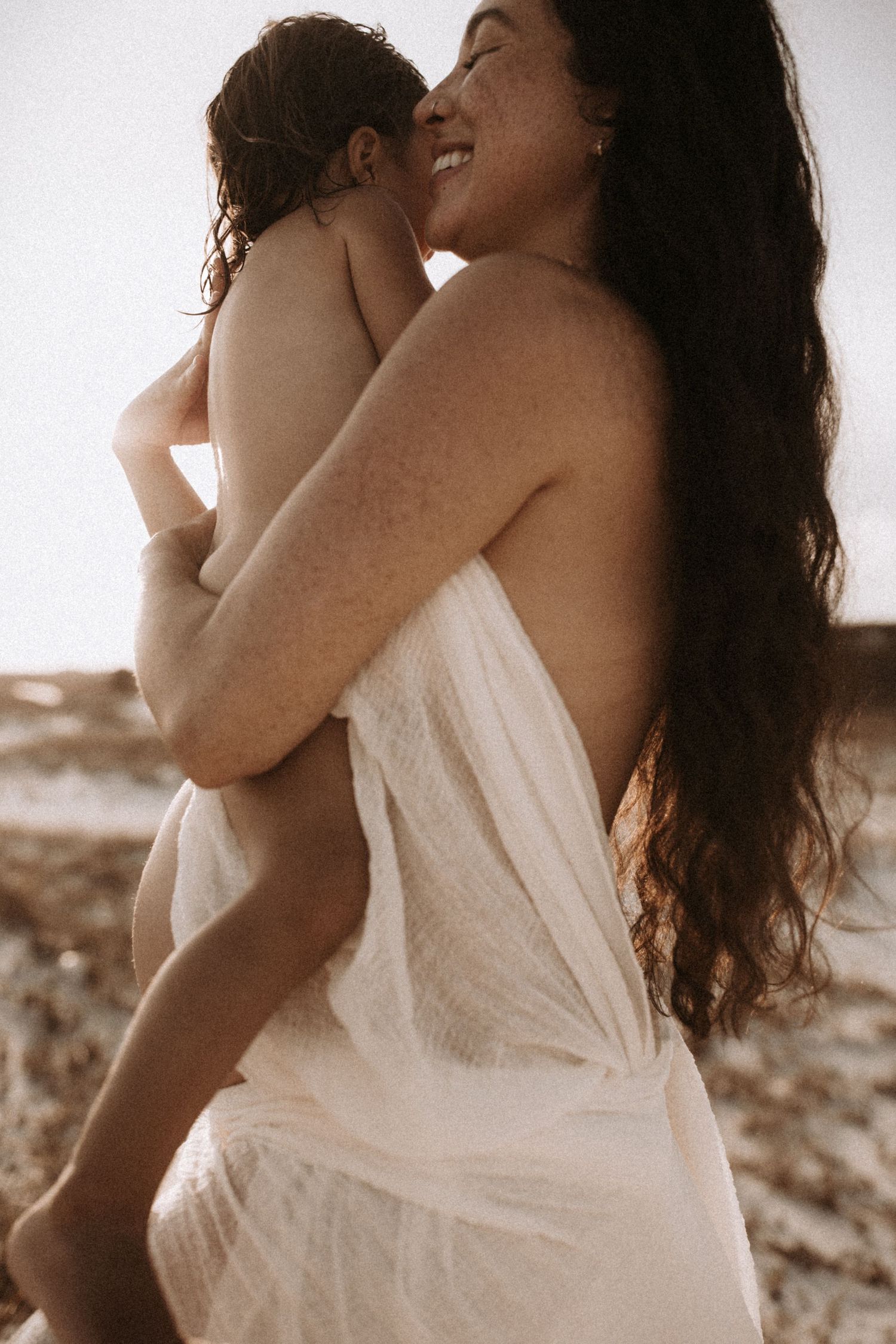 Pregnant woman with freckles and long dark hair smiles while holding young child on beach.