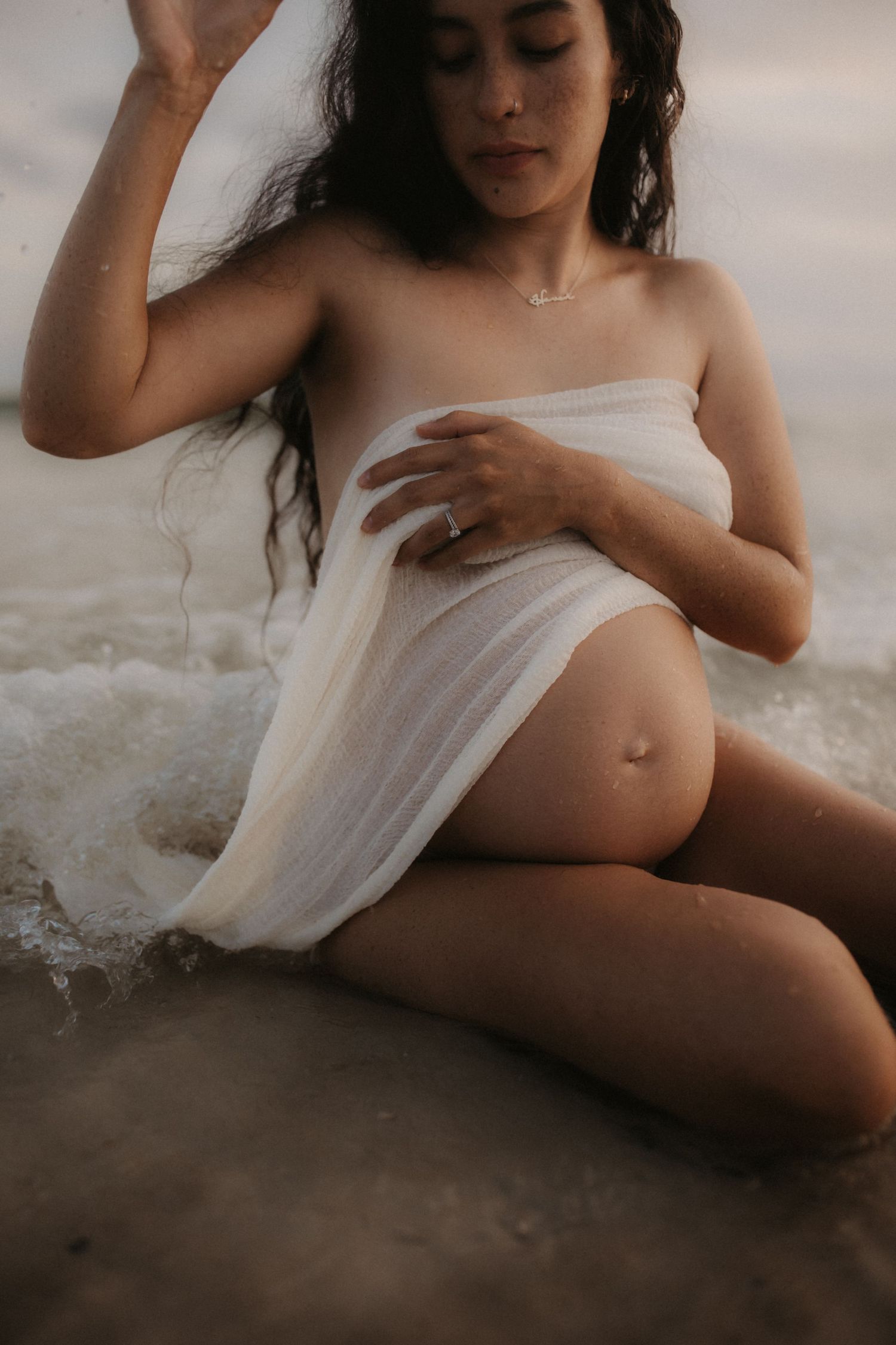 A pregnant woman wrapped in white sheer fabric sits gracefully on a beach with ocean waves around her.