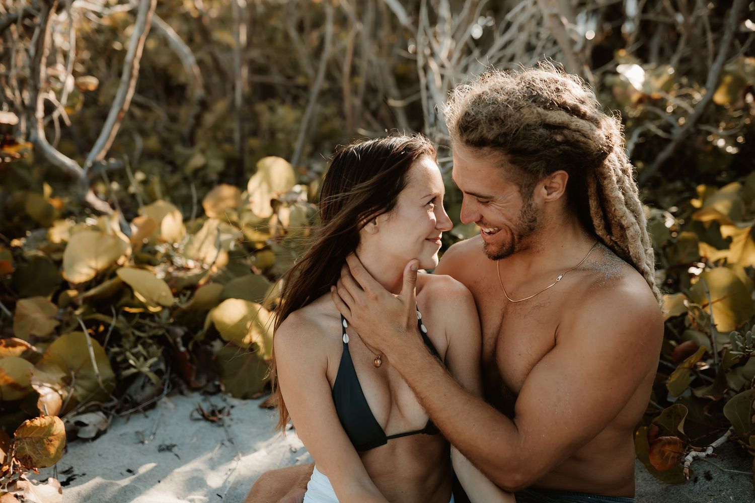 Romantic couple with dreadlocks embracing among tropical foliage in sunlight.