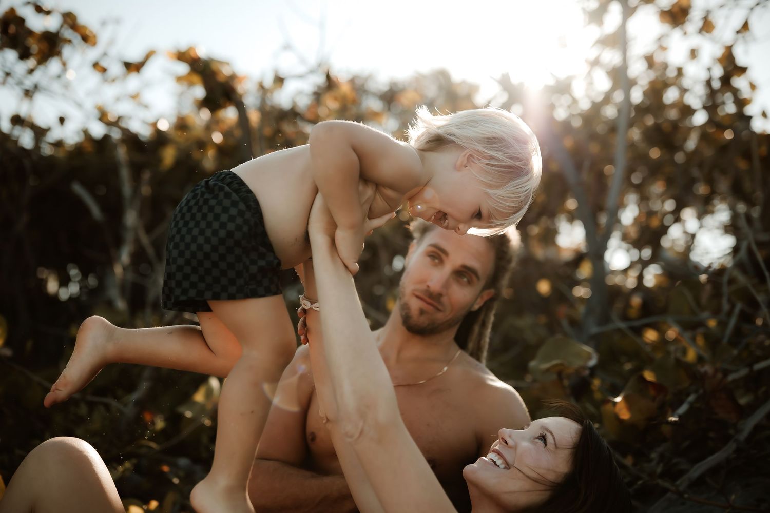 Happy couple lifting a toddler in the air together outdoors surrounded by lush greenery.