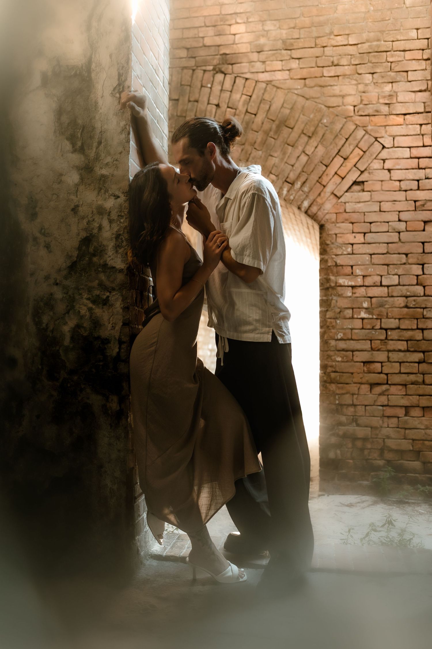 A couple shares a romantic moment dancing in a rustic brick archway, bathed in soft dramatic lighting.