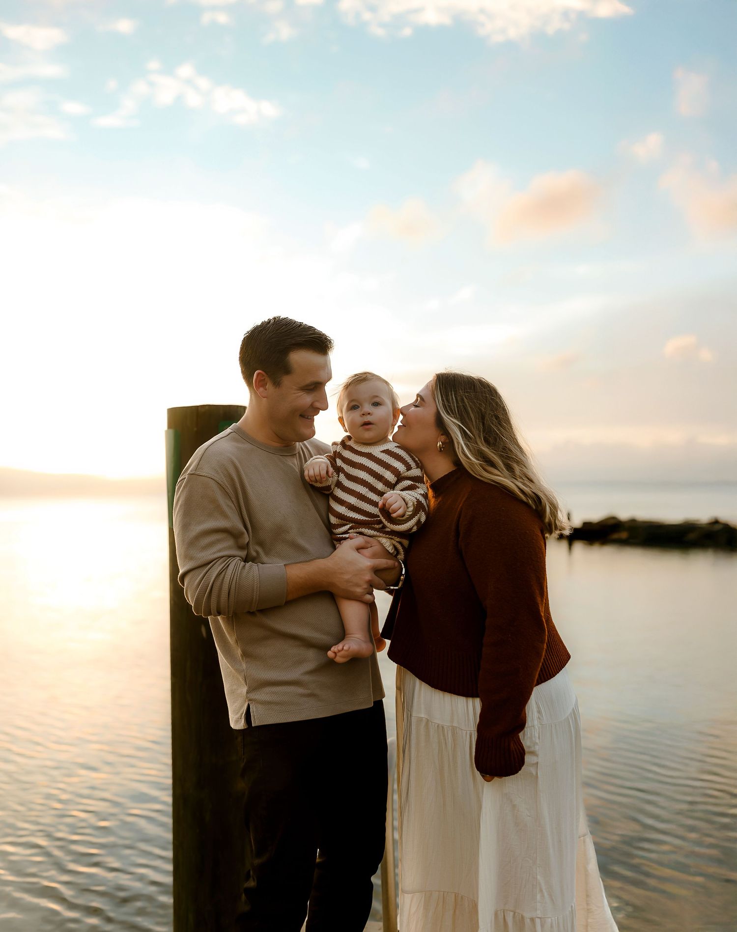 A couple shares a tender moment with their baby by a waterfront dock at golden hour sunset.