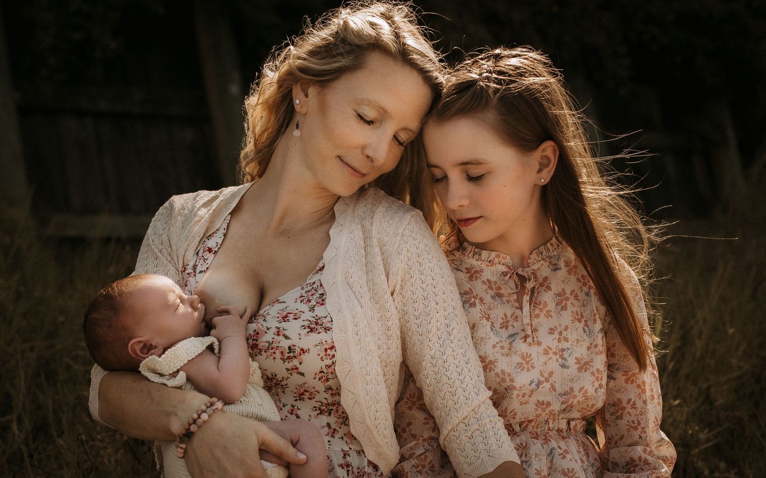 Mother with newborn baby and young daughter in matching floral outfits outdoors.
