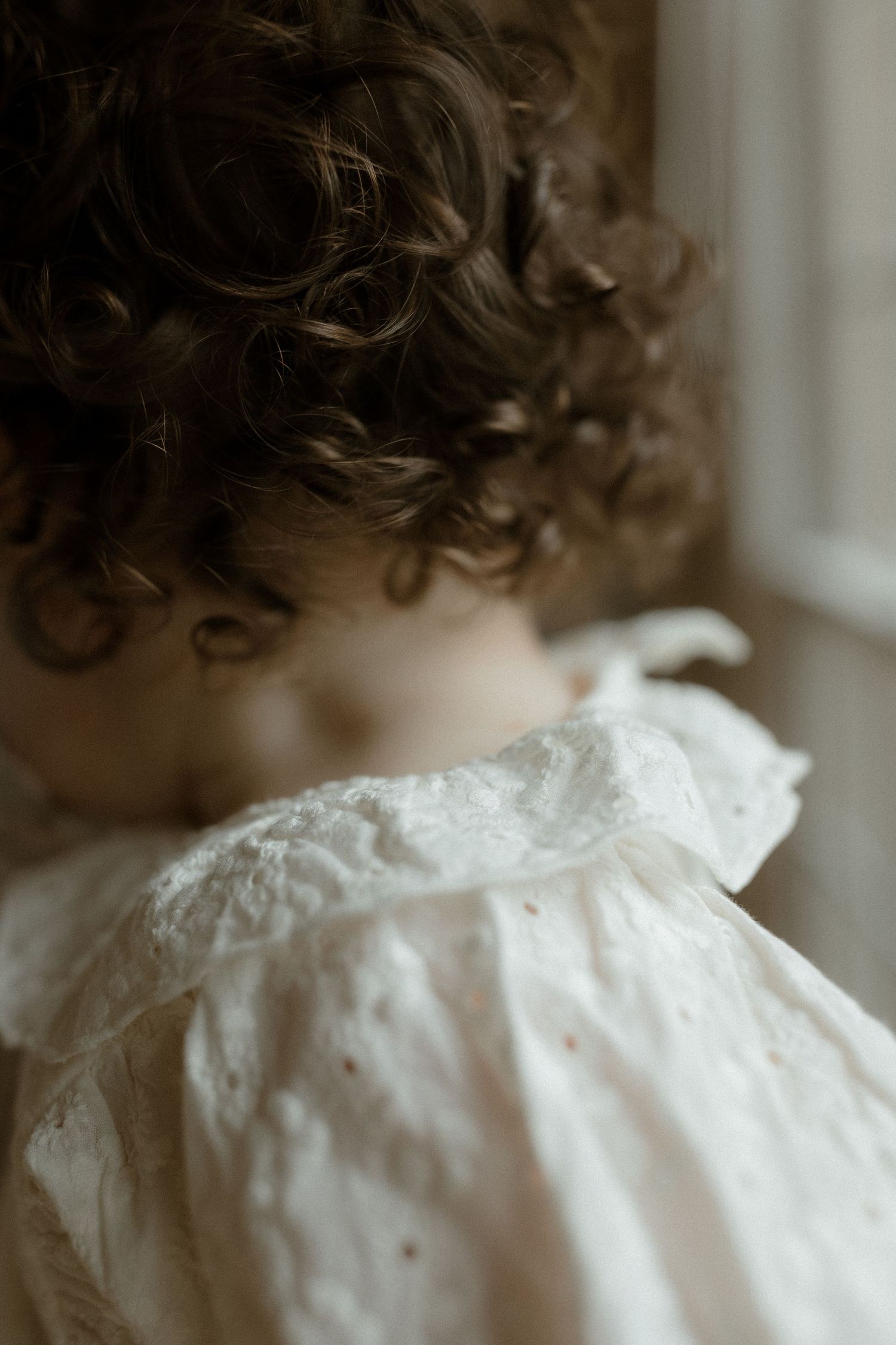 Close-up of a young child with curly brown hair wearing a white lace dress, gazing out a window.