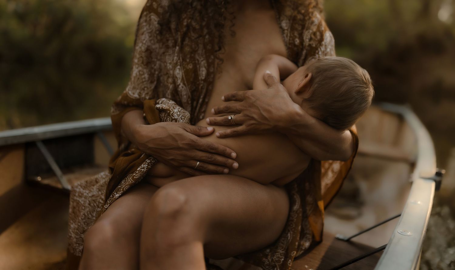 A mother breastfeeds her infant outdoors near a wooden boat in a natural forest setting.