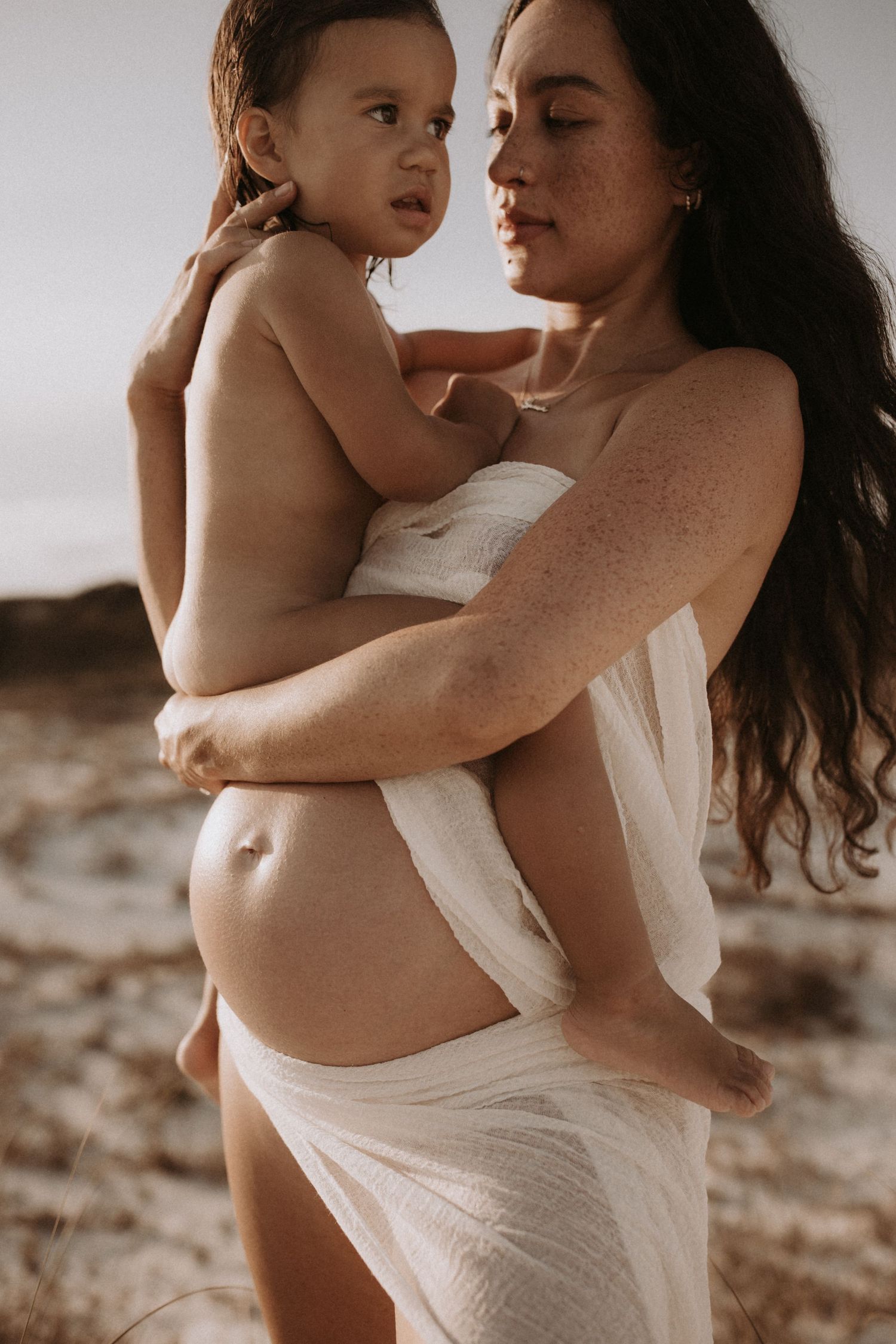 Pregnant freckled mother holding toddler on sandy beach, wrapped in white fabric.
