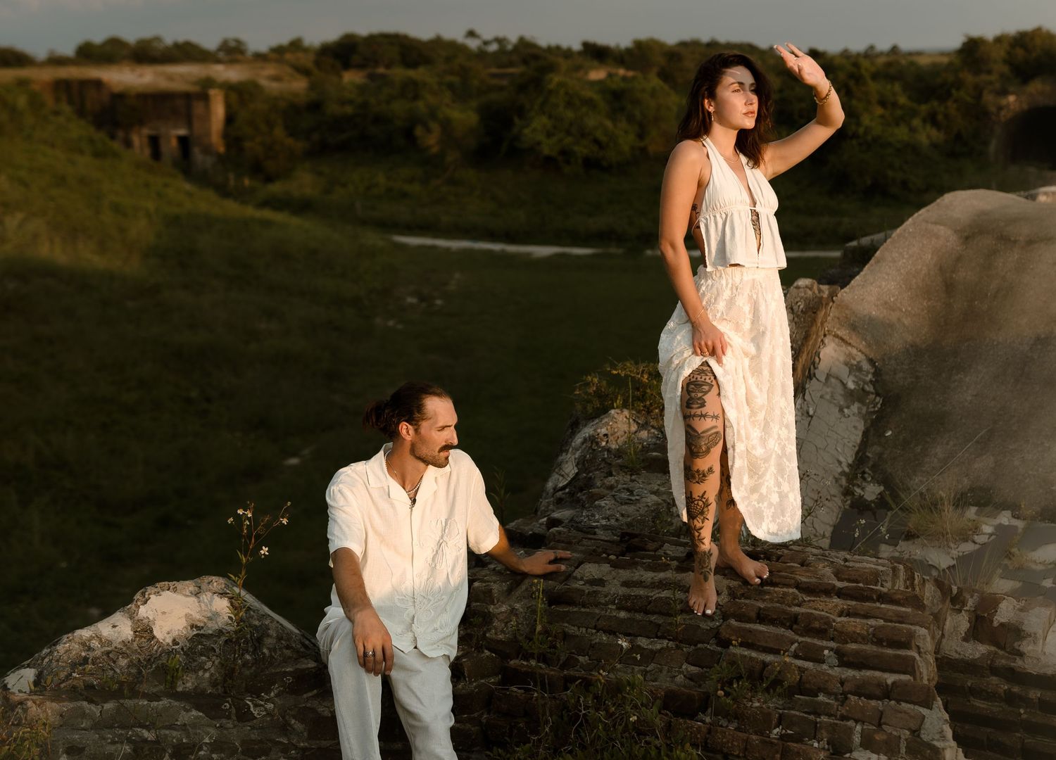 A man and woman in white outfits pose on ancient stone ruins in a sunlit green landscape.