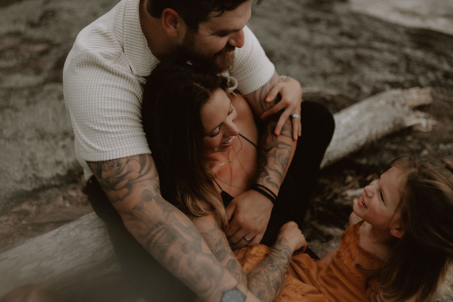 A tattooed man embraces a woman and child in a warm, candid family photography session outdoors.