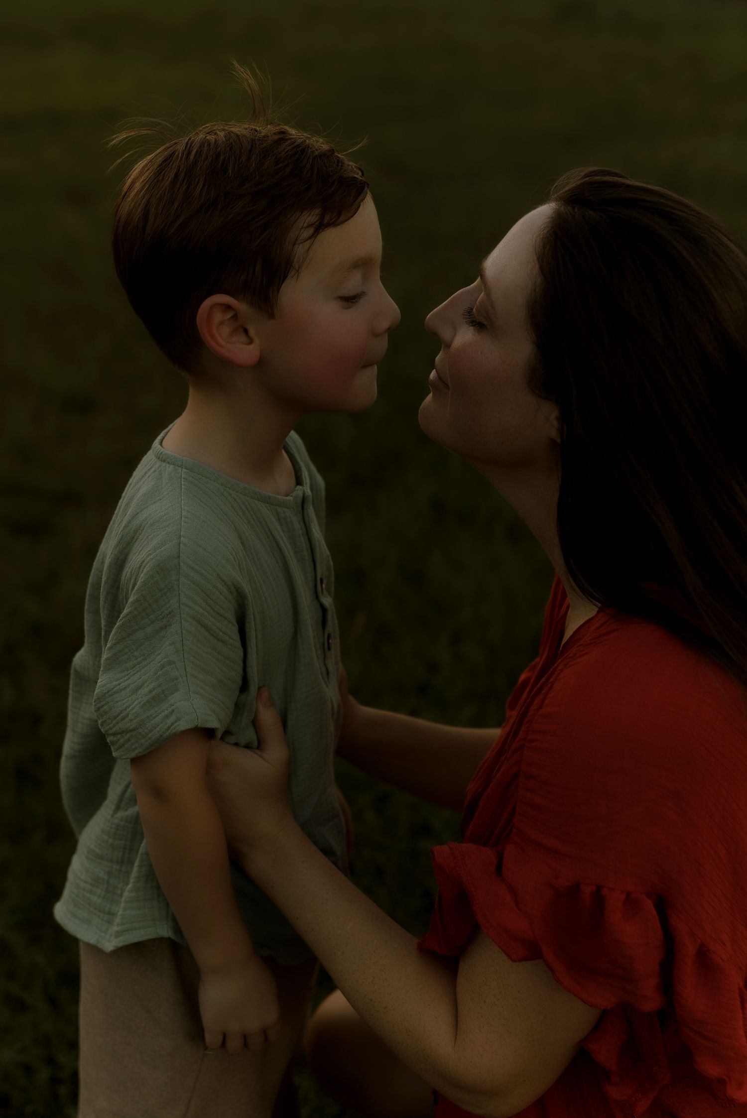 A mother in a red dress tenderly holds her young son close as they share a sweet nose-to-nose moment outdoors.