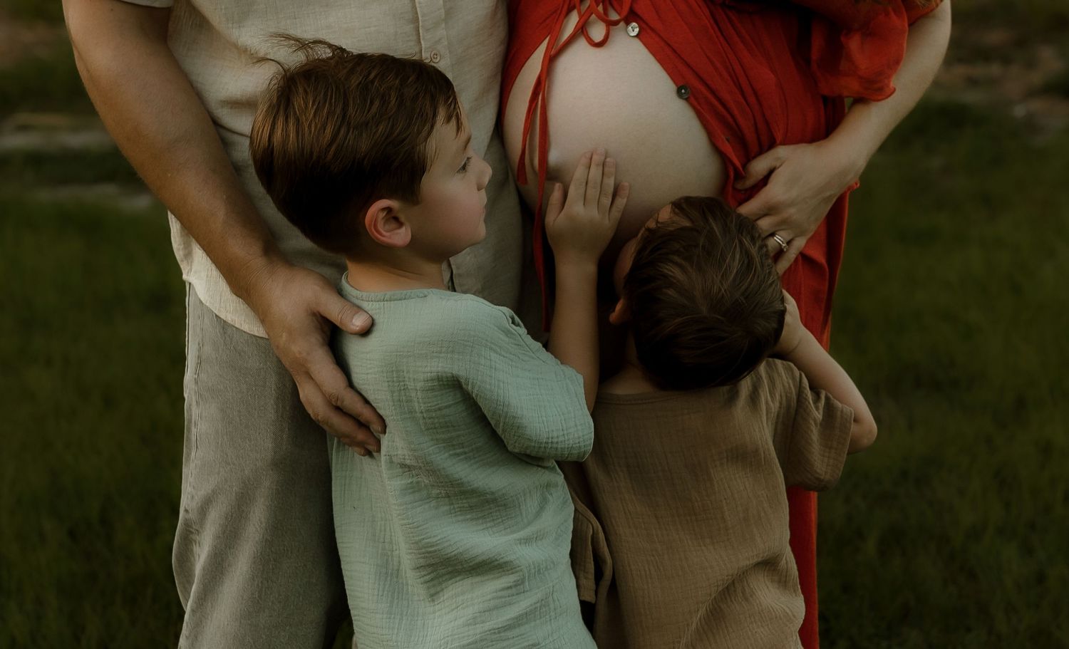 Pregnant woman in red dress with two young children hugging her baby bump outdoors in a field.
