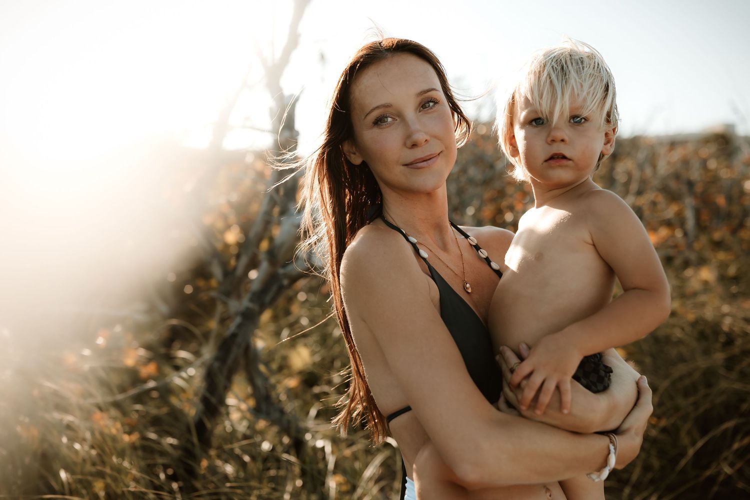 A mother holds her young toddler outdoors in golden sunlight among tall dry grasses.