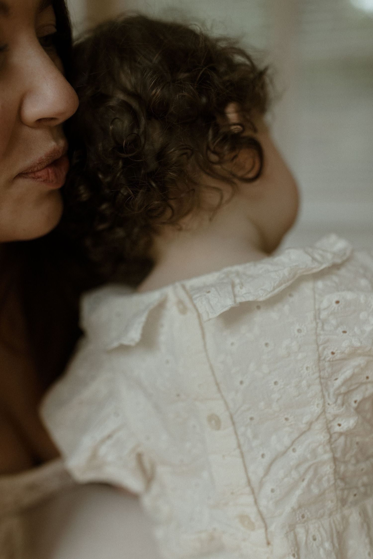 A mother with curly hair holds her baby dressed in a white eyelet lace outfit, captured in soft natural light.