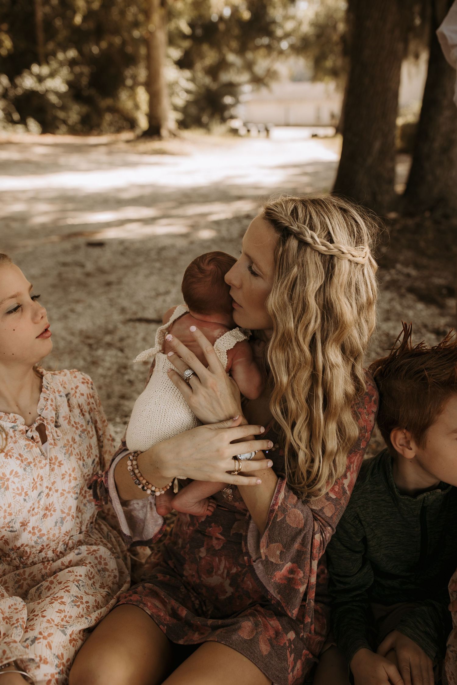 A mother with braided hair kisses her newborn baby while sitting outdoors with two other children nearby.