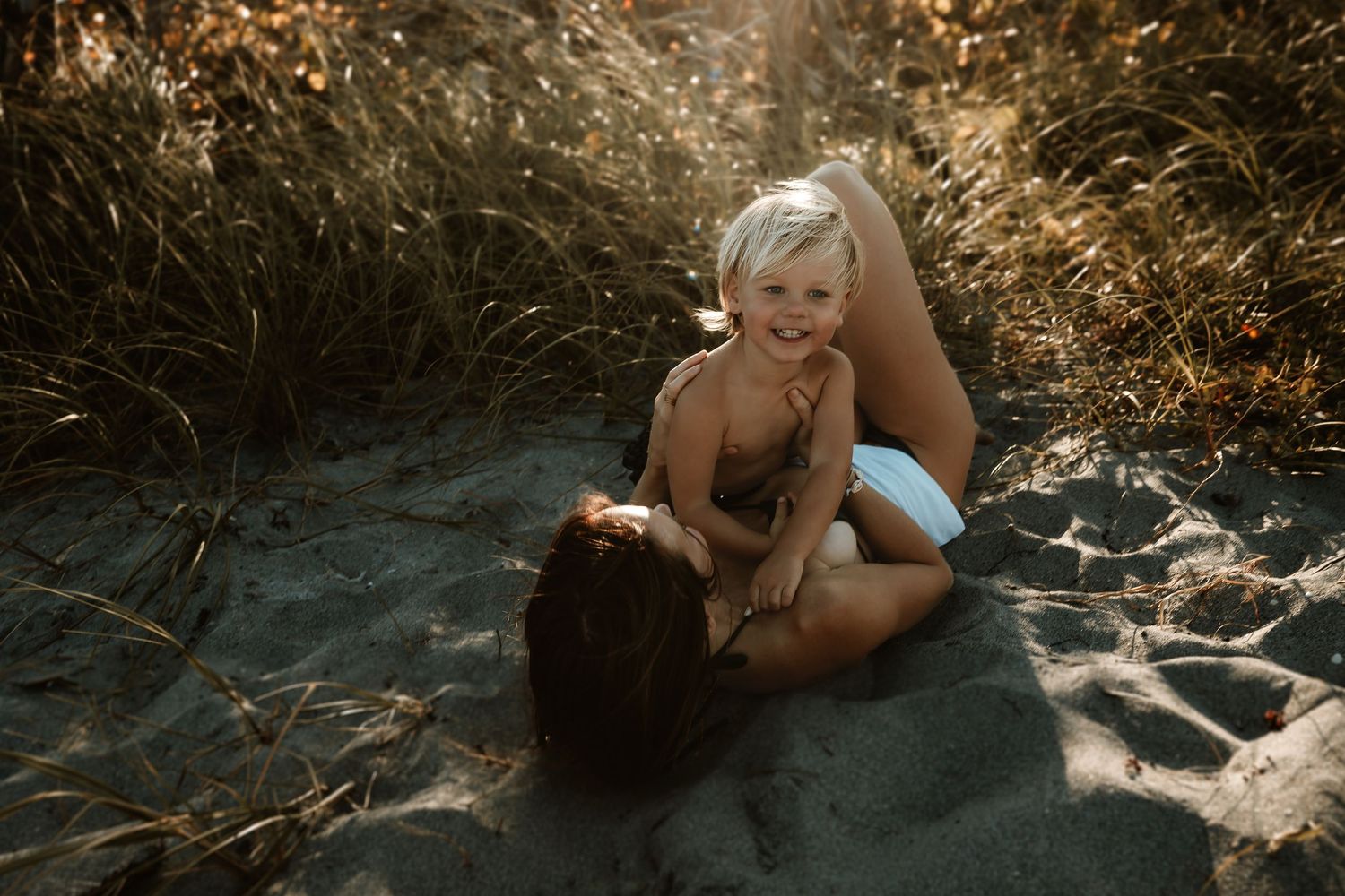 Laughing blonde toddler playing with mother on sandy beach surrounded by tall grass.