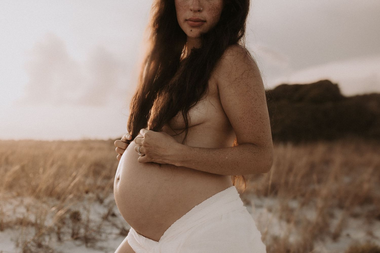 Pregnant woman in white skirt holding baby bump outdoors in golden hour light on sandy beach.