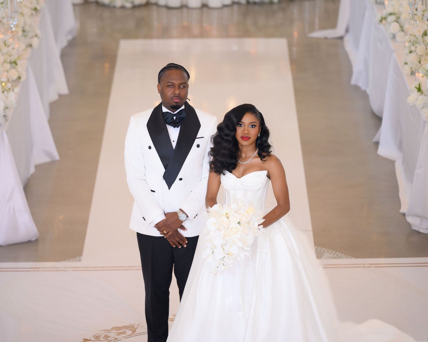 Elegant wedding couple poses at altar, groom in white tuxedo, bride in strapless ball gown holding white bouquet.