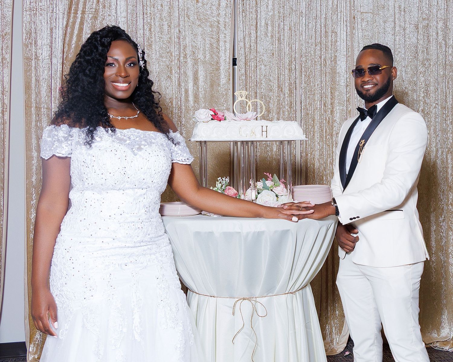 Bride and groom in elegant white attire standing beside a tiered wedding cake at their reception.