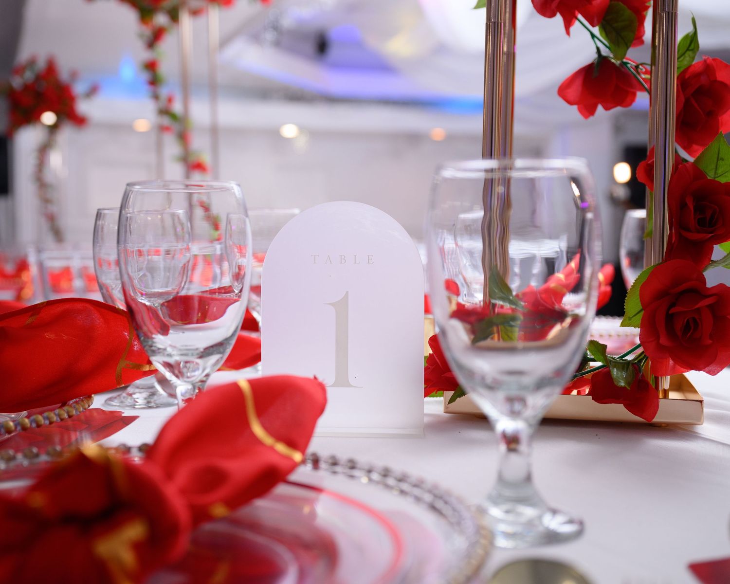 Elegant wedding table setting with red roses, crystal glasses, and a white table number card on a white tablecloth.