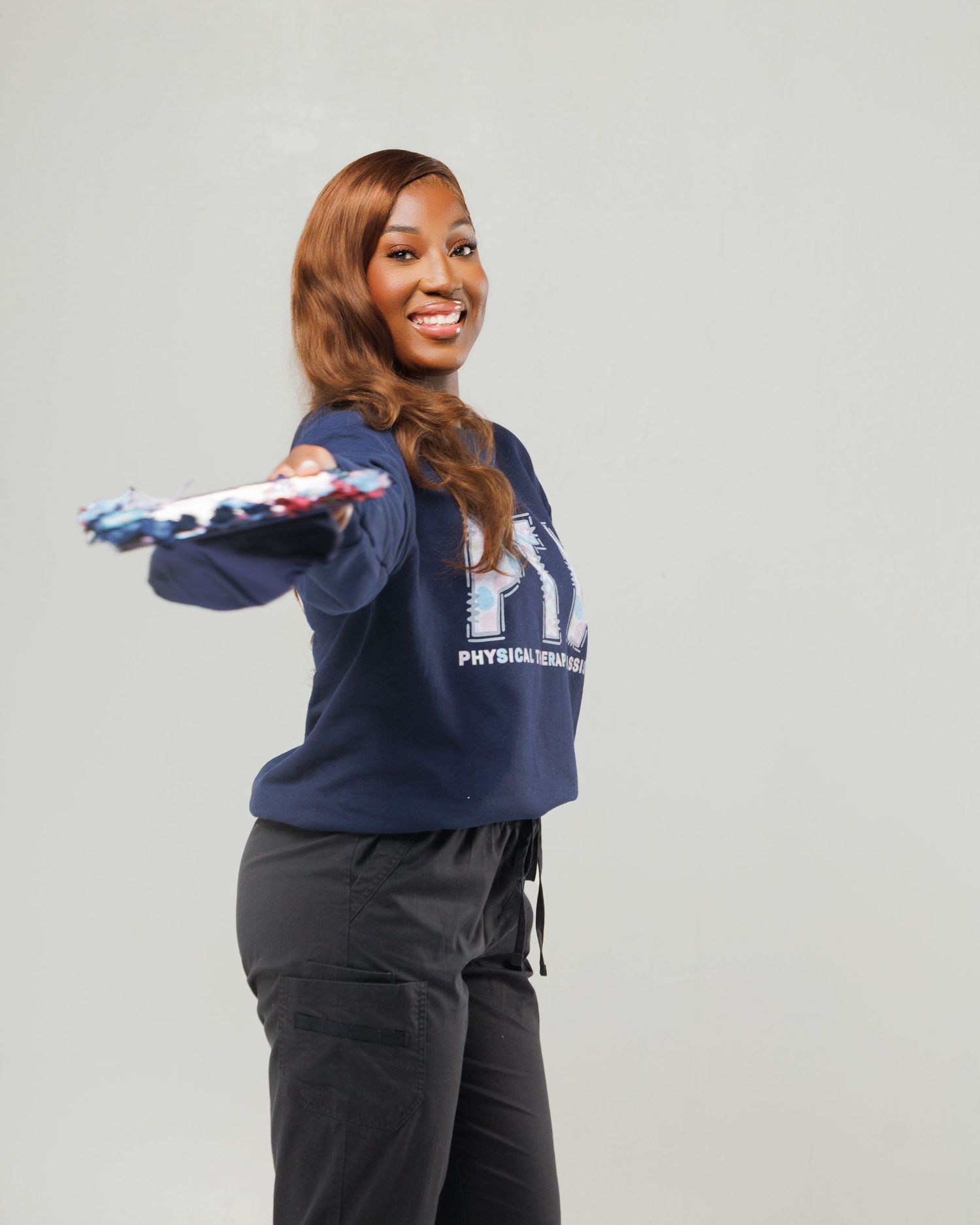 Smiling woman in navy Physical Therapy sweatshirt and scrubs extends American flag toward camera.