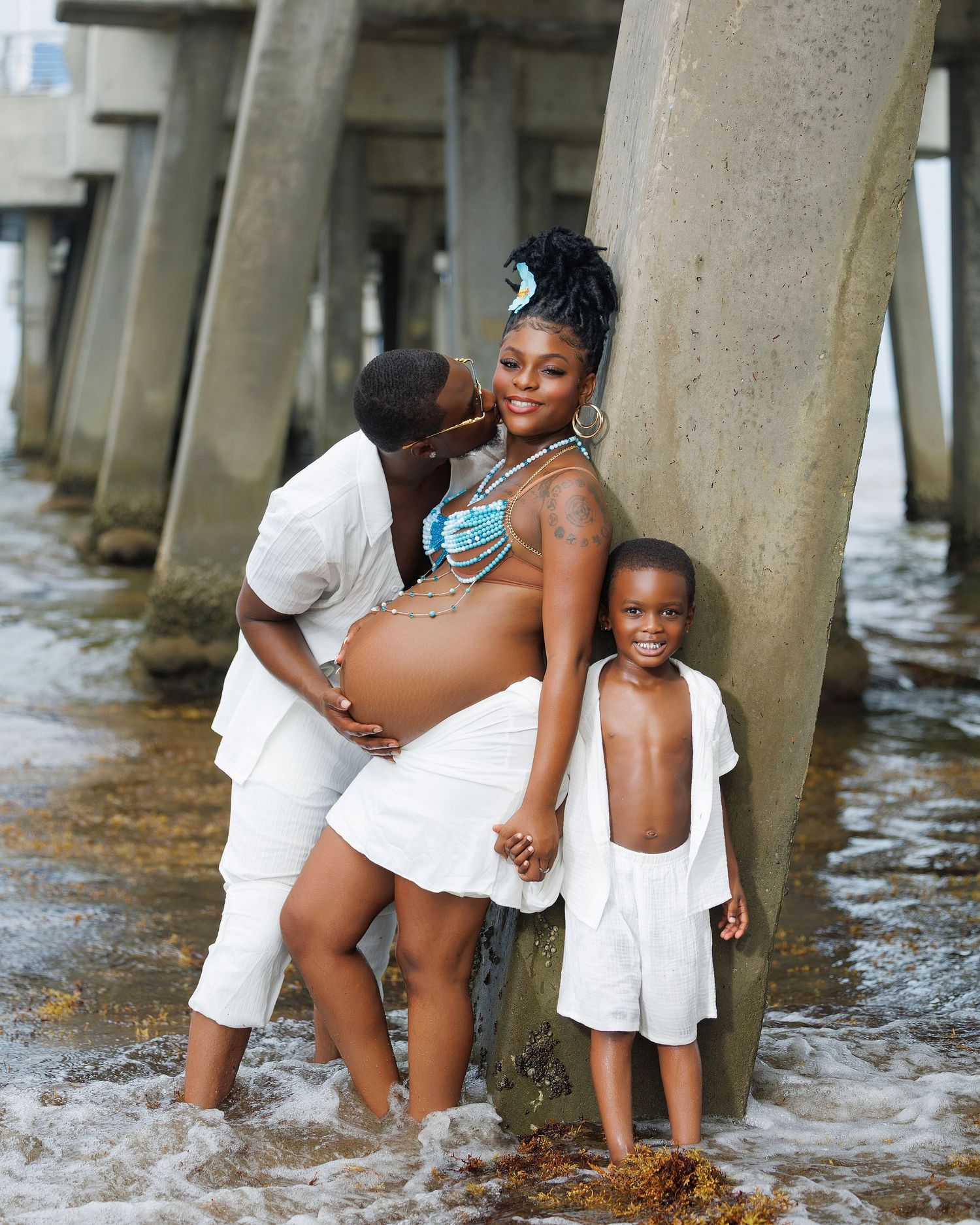 Pregnant woman in blue beaded top poses with partner and child under a pier at the beach.