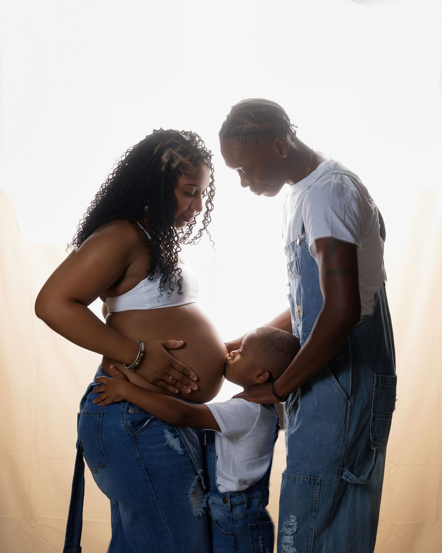 Pregnant Black woman with curly hair and her partner in denim overalls share a tender family moment with their young child.