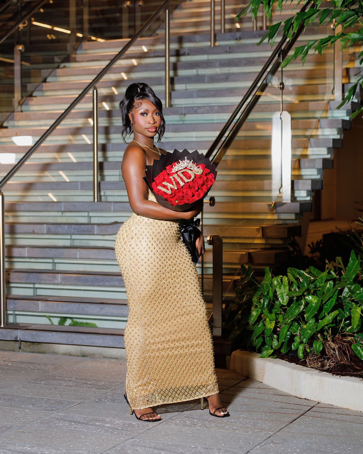 Woman in gold skirt and black top holds red rose heart bouquet at elegant venue staircase at night.