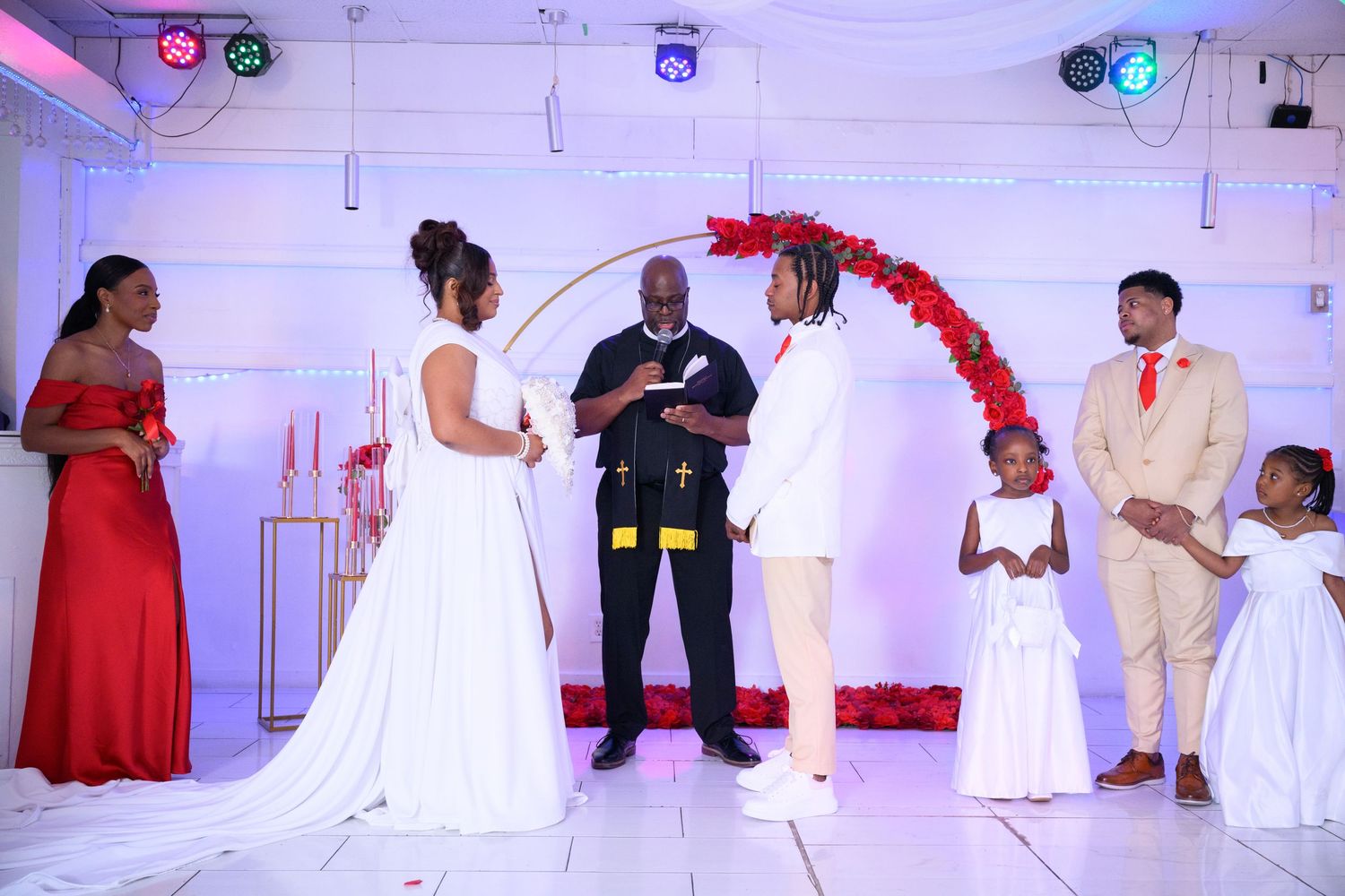 Wedding ceremony with bride and groom exchanging vows before officiant, flanked by guests in red and cream attire.