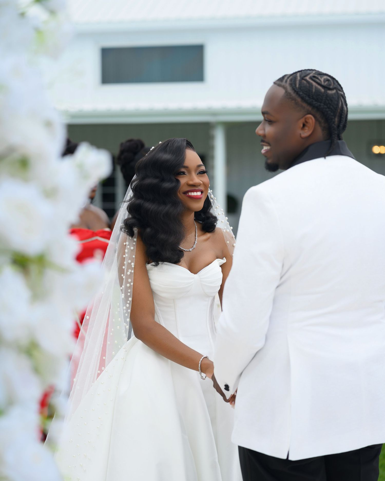 A bride in a white strapless gown and veil smiles at her groom in a white tuxedo jacket during an outdoor wedding ceremony.