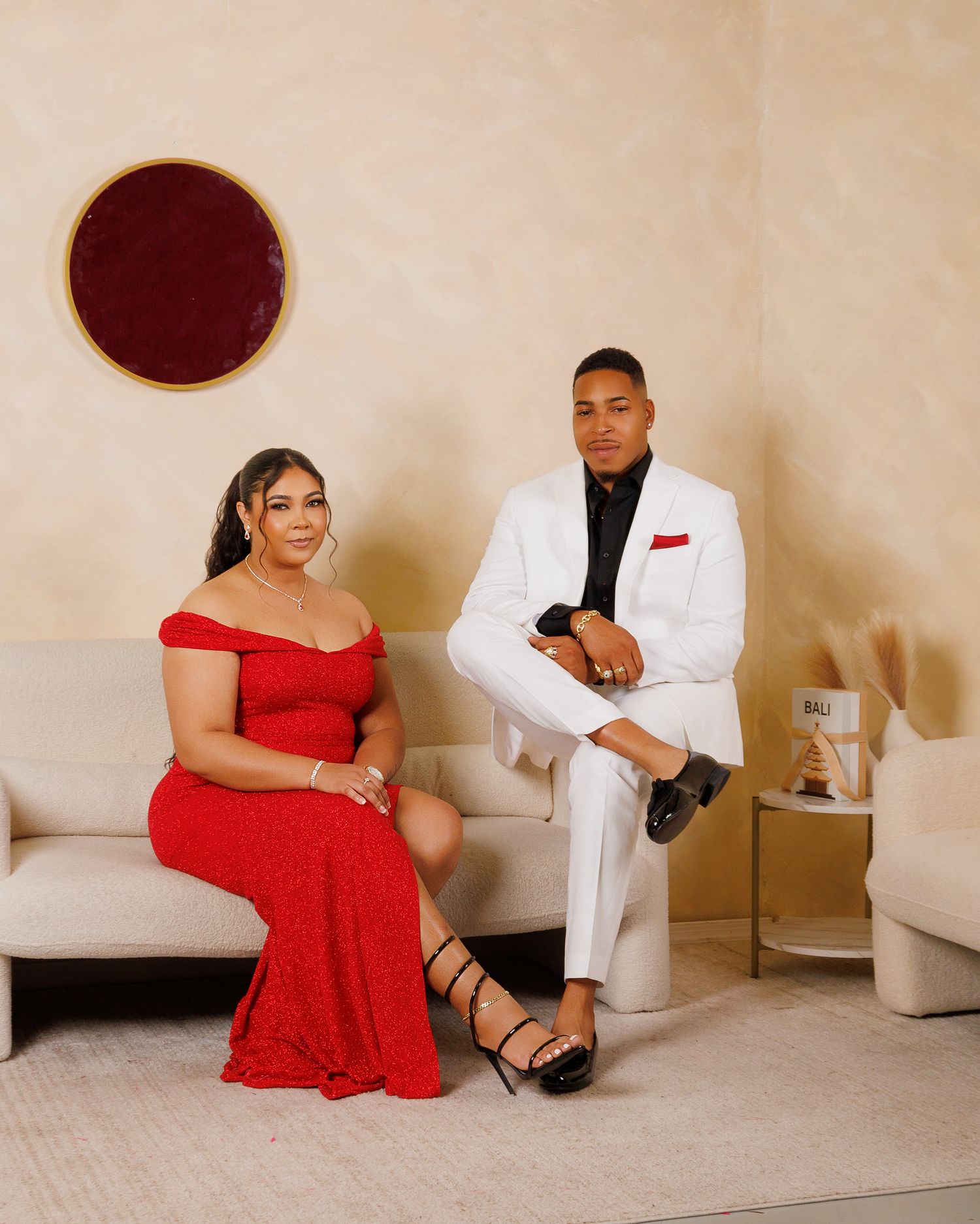 A woman in a red off-shoulder gown and a man in a white suit with red pocket square pose elegantly on a cream sofa.