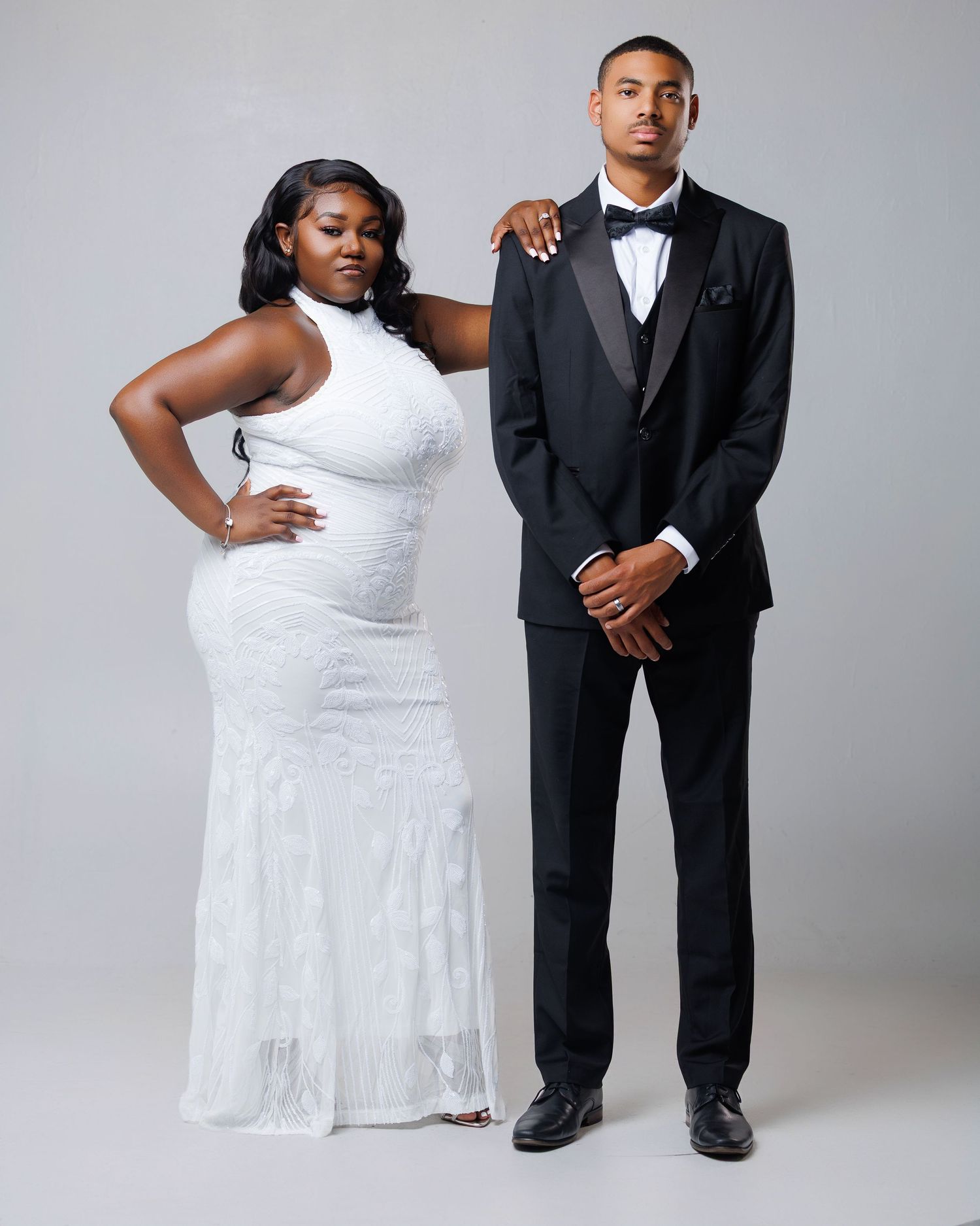 Couple posing formally, woman in white halter gown and man in black tuxedo with bow tie, against gray backdrop.