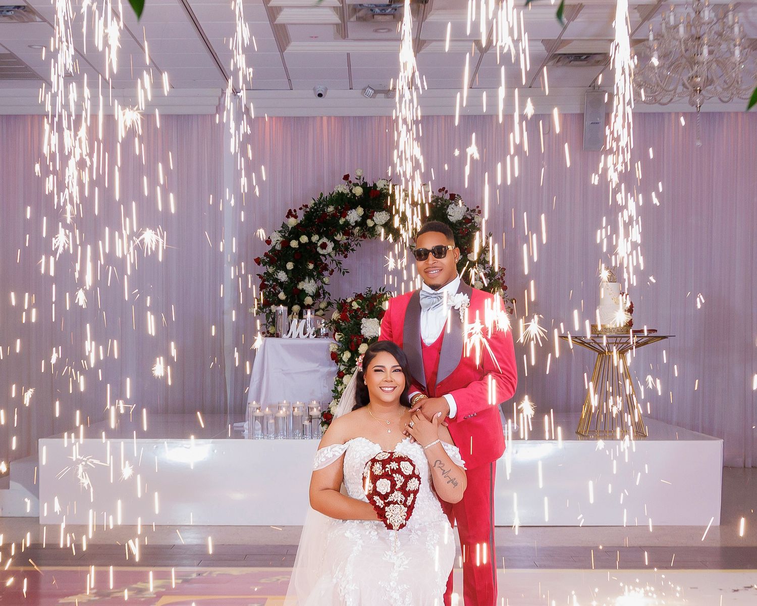 Bride and groom posing at wedding reception with sparkler fountains, bride in white gown holding red bouquet, groom in red suit.