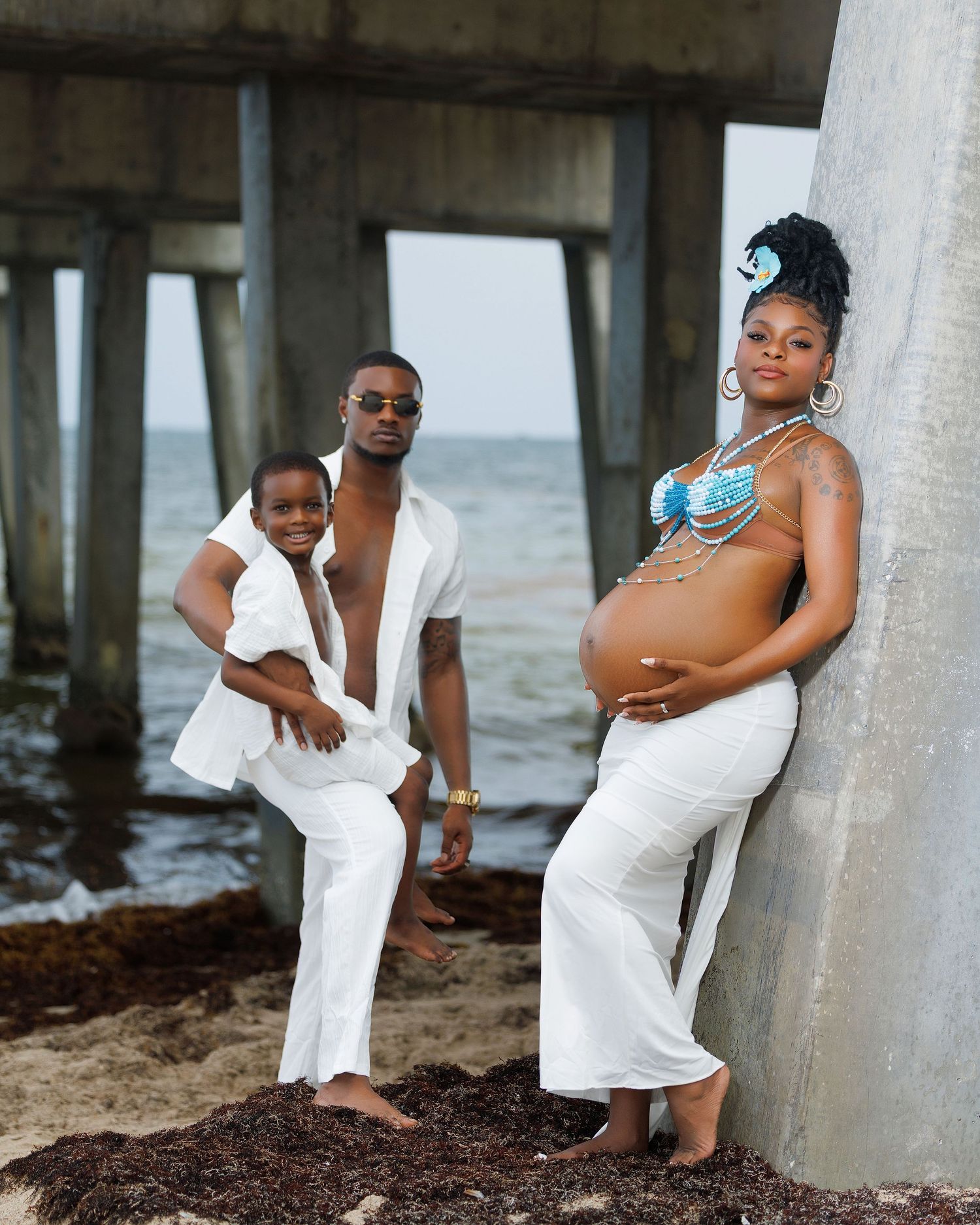 A pregnant woman in a blue bikini top and white pants poses with her partner and child by a beach pier.