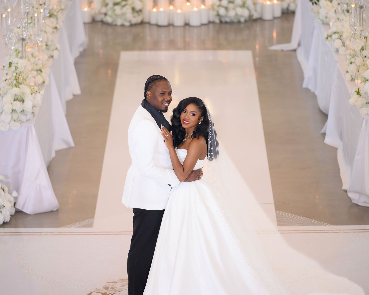 A bride and groom pose elegantly in a white wedding venue lined with tall floral centerpieces.