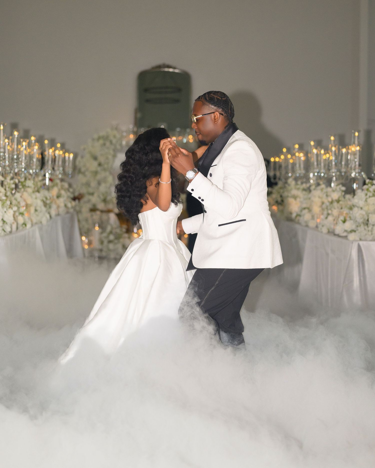 Bride and groom share first dance surrounded by elegant fog, candles, and white floral arrangements at their wedding reception.