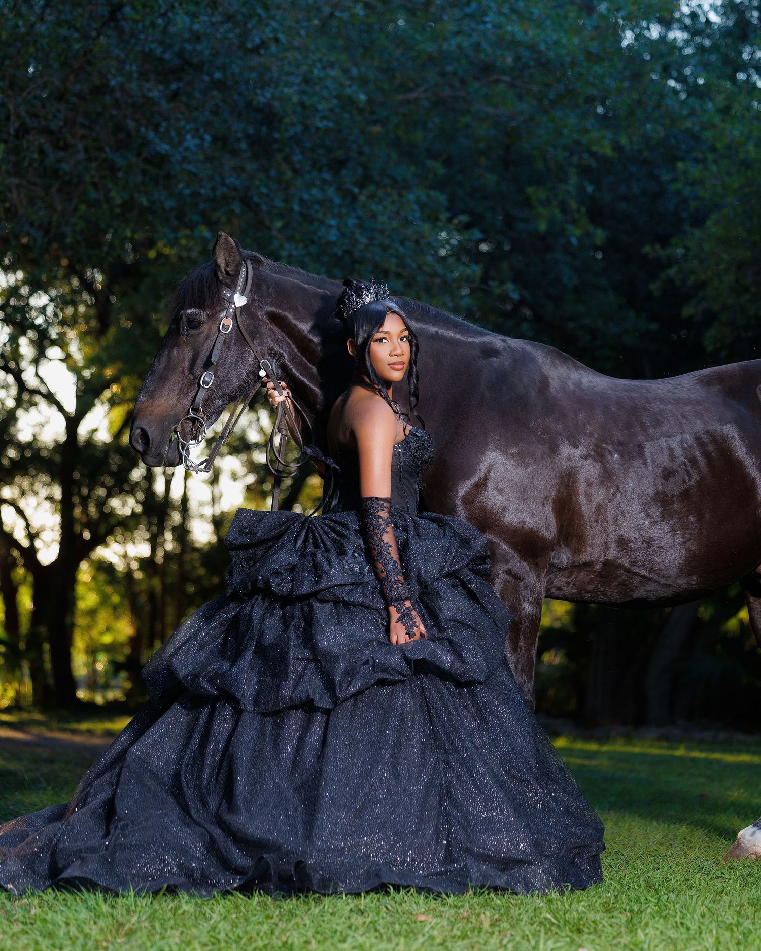 A young woman in a stunning black ball gown poses elegantly beside a dark horse in a lush green outdoor setting.