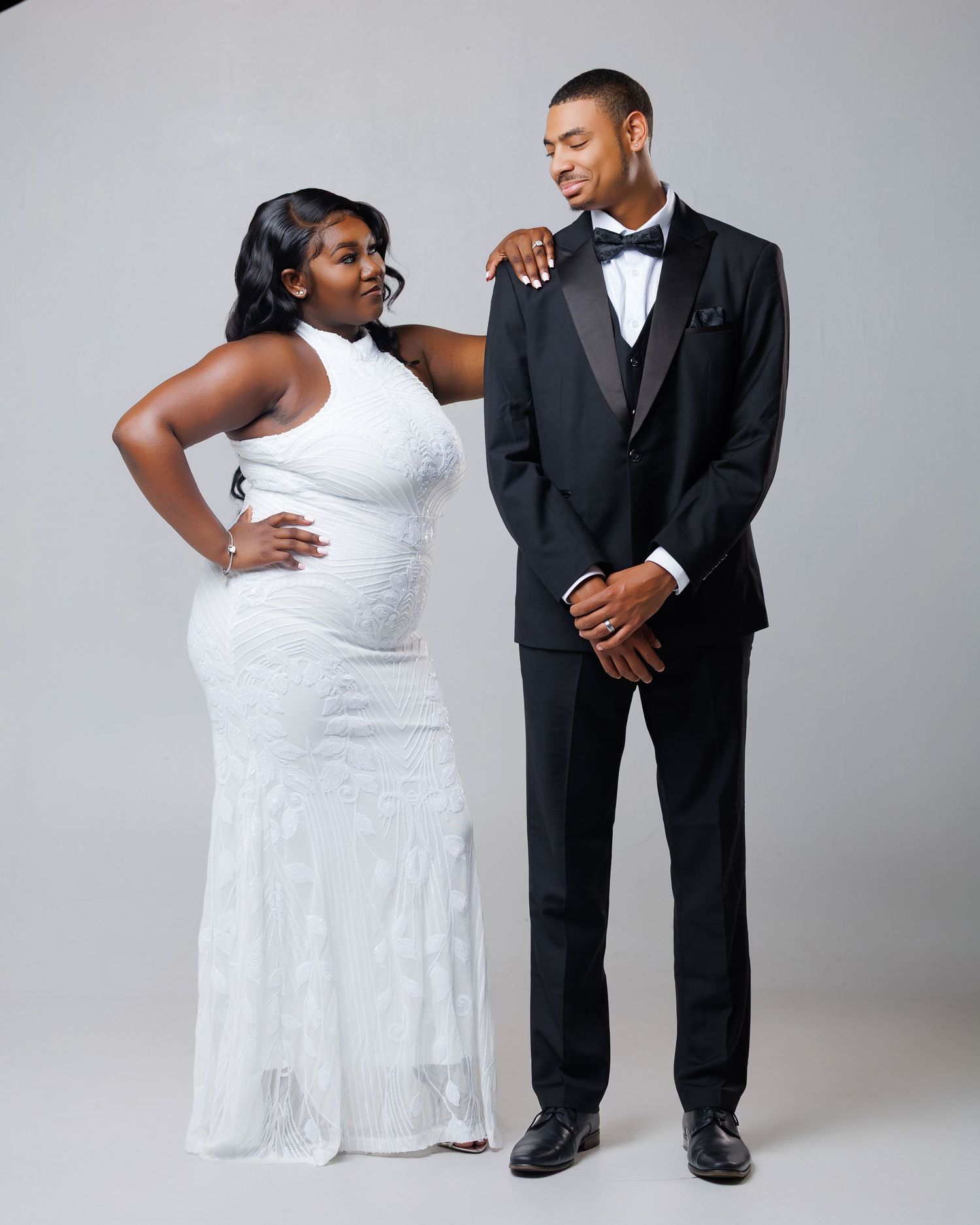 Couple posing in formal wear, woman in white halter gown and man in black tuxedo with bow tie.