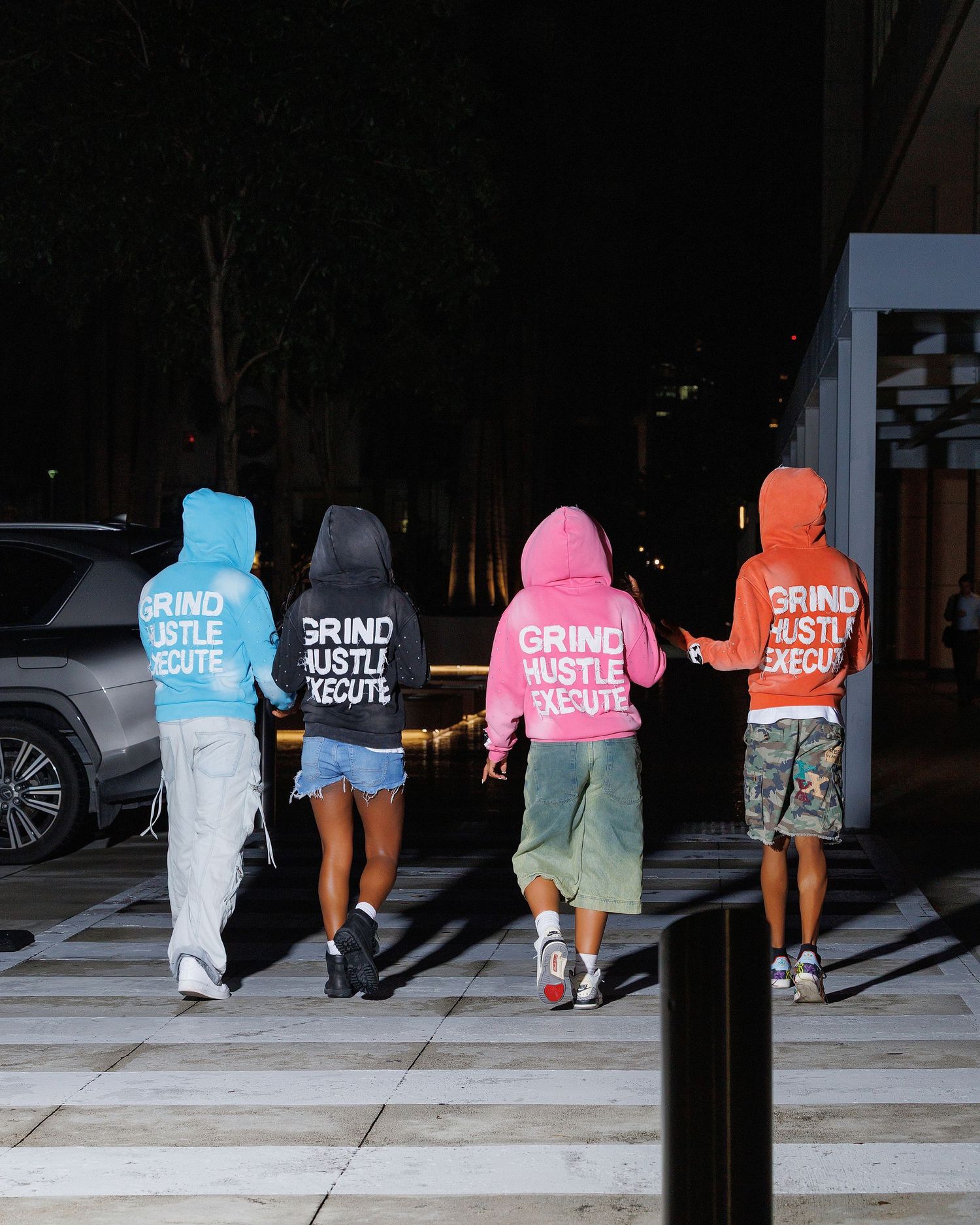 Four people walking away wearing colorful Grind Hustle Execute hoodies in blue, gray, pink, and orange at night.