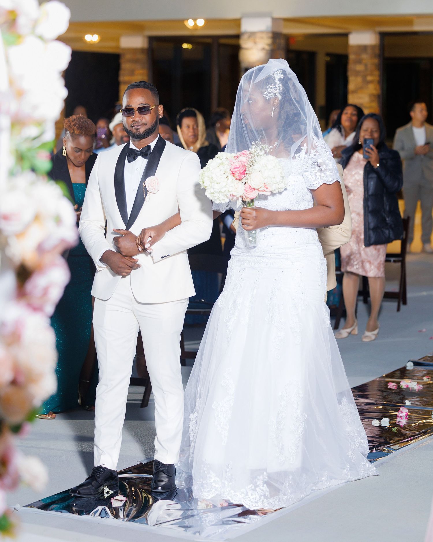 Elegant wedding ceremony with groom in white tuxedo and bride in lace gown holding pink bouquet walking down the aisle.