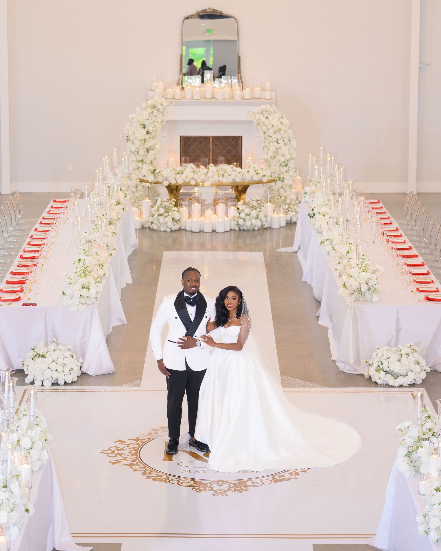 Elegant wedding reception with couple posing in white floral-decorated ballroom with candles and long banquet tables.