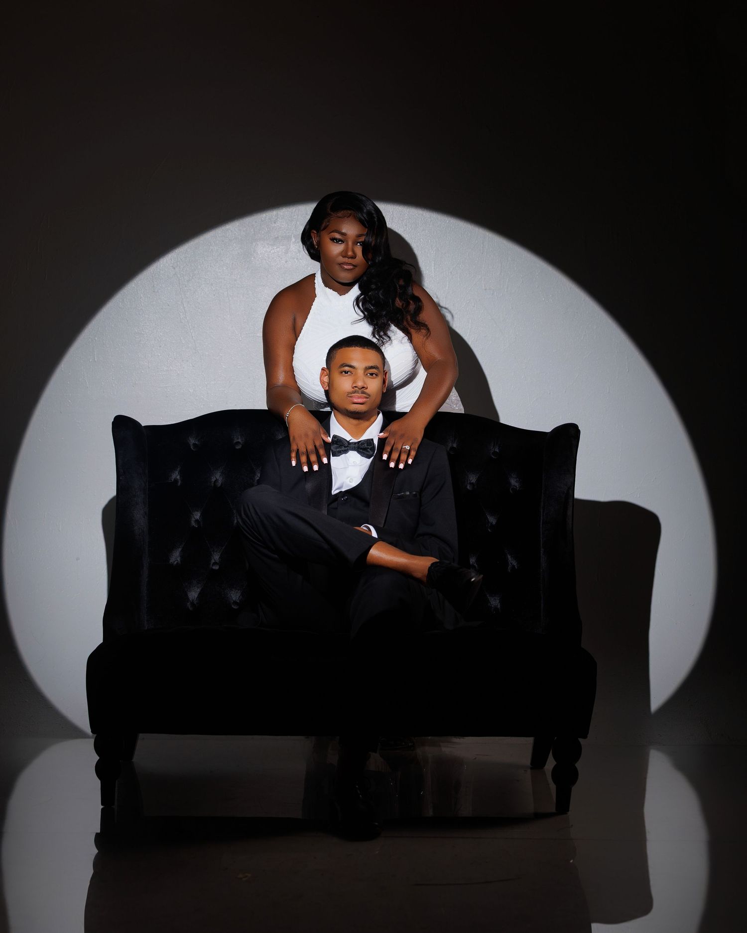 A couple in formal black and white attire poses elegantly on a tufted black sofa with dramatic spotlight backdrop.