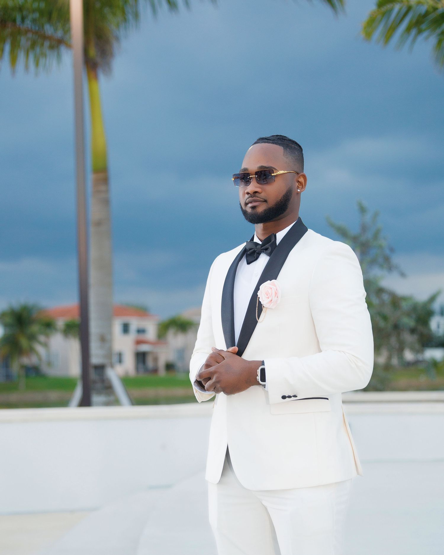 Elegant man in white tuxedo with black lapels and bow tie posing outdoors at a tropical wedding venue.