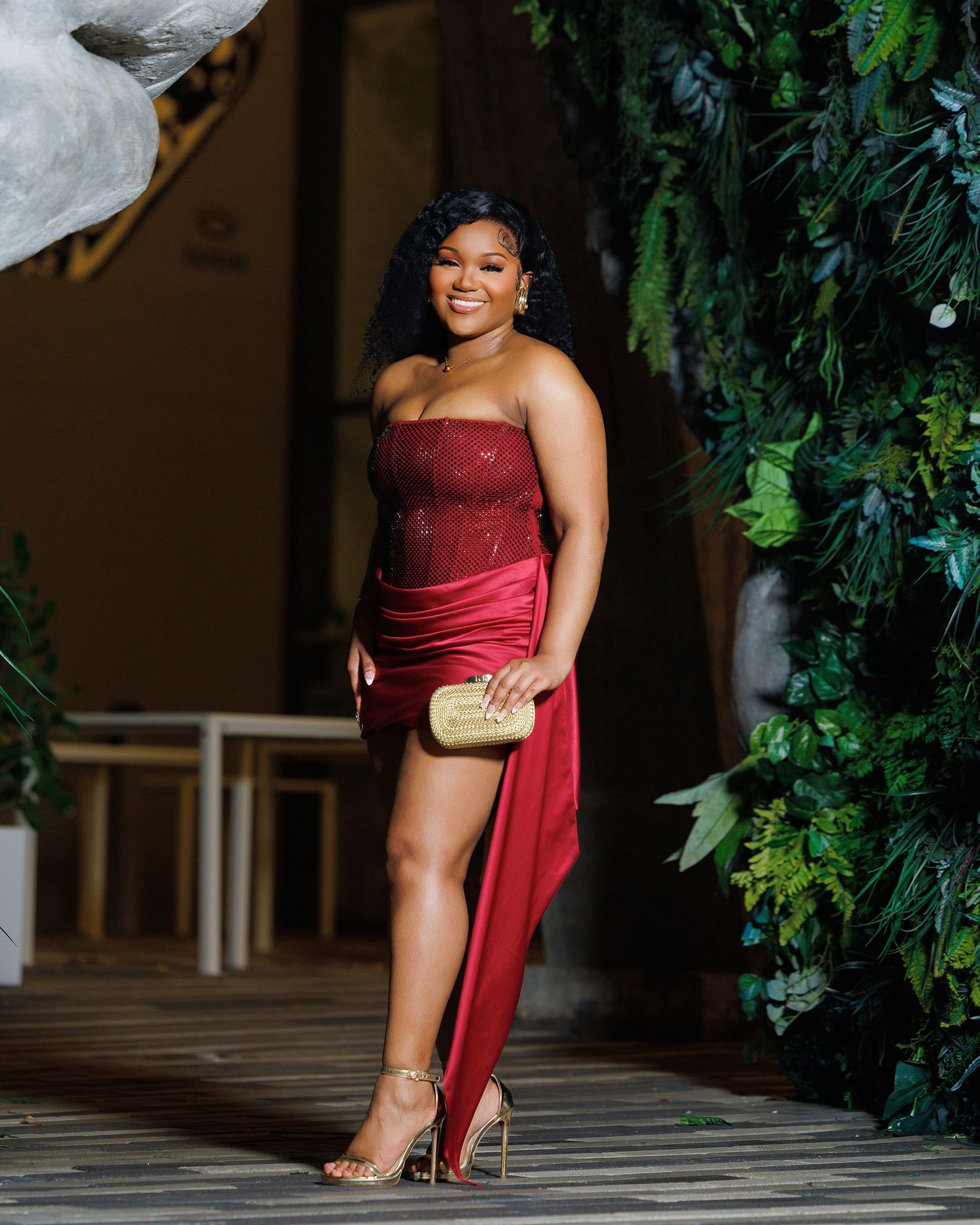 Woman in elegant red strapless dress with high slit, gold heels and clutch, posing near lush green wall.