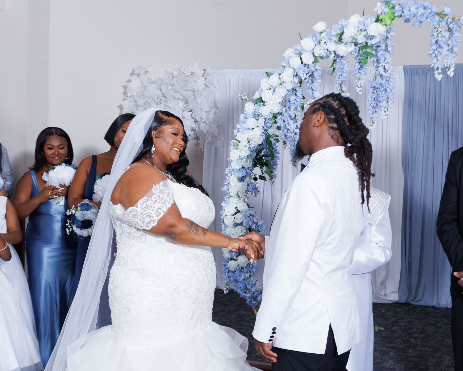 Bride and groom holding hands at wedding altar with blue floral arch and bridesmaids in blue dresses.
