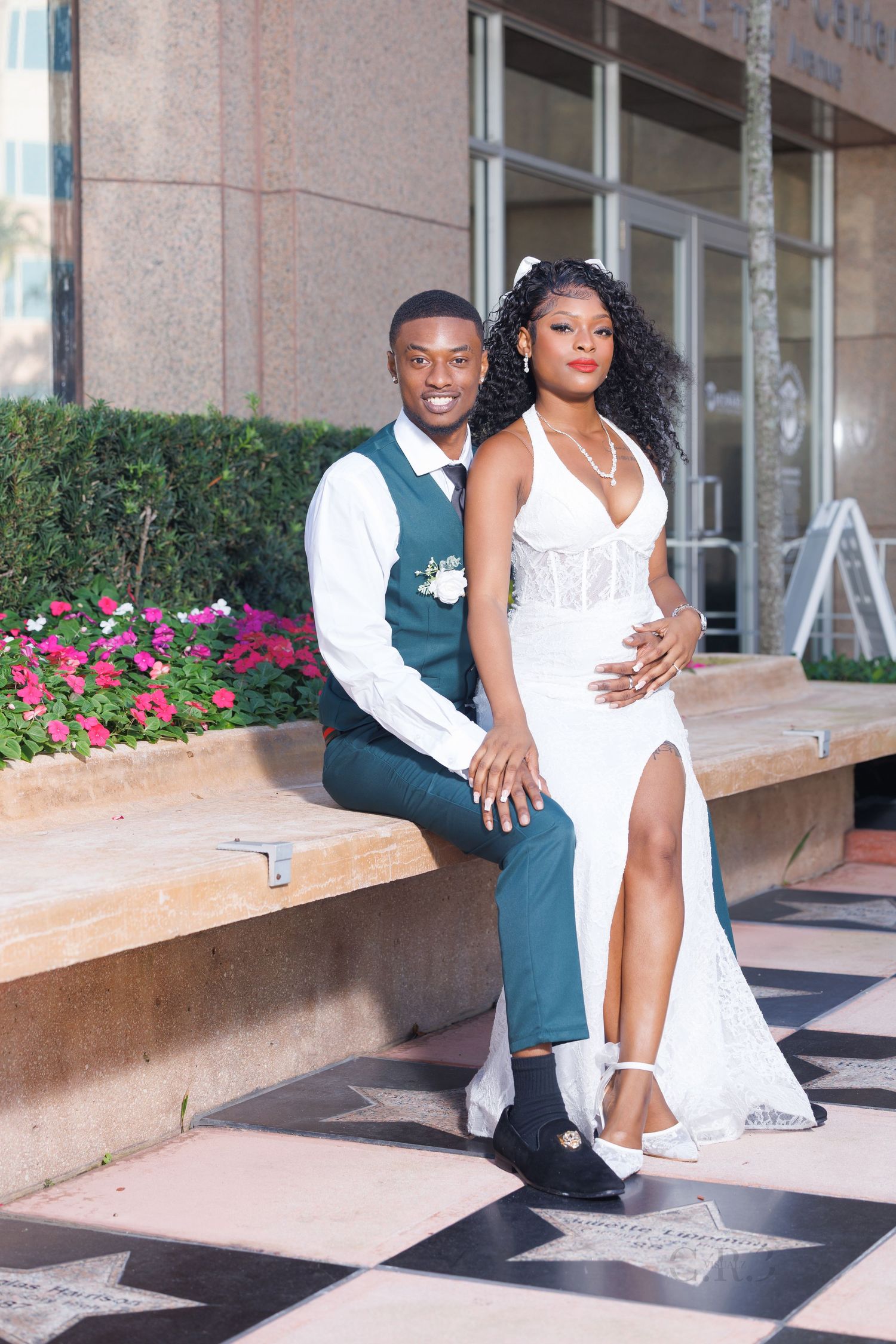 Elegant newlyweds pose on a bench outdoors, groom in teal vest and bride in white lace gown with slit dress.