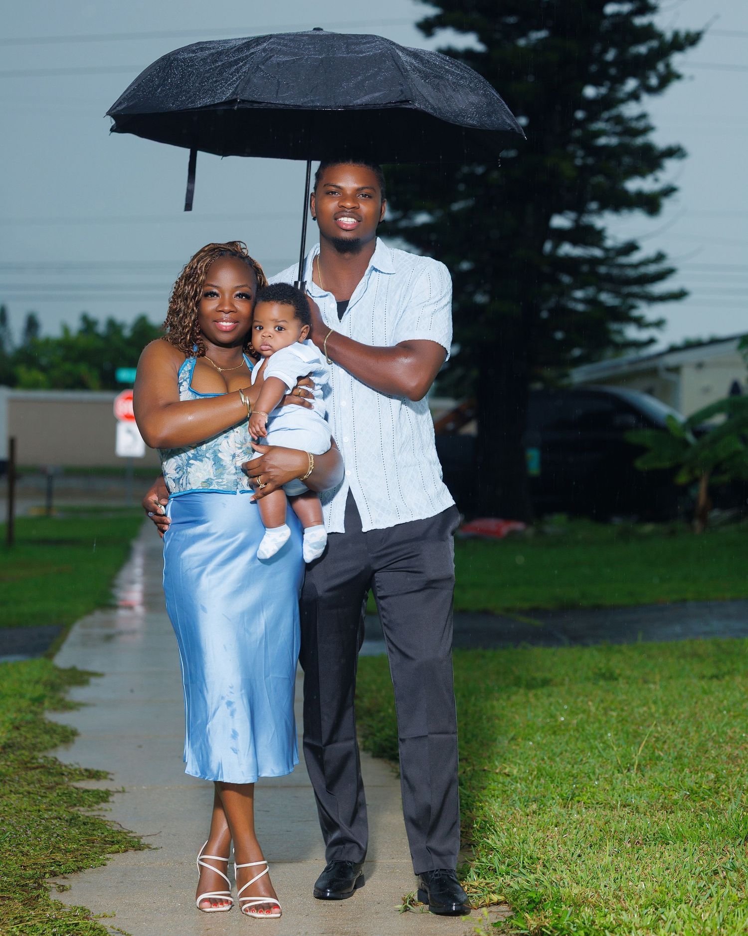 A well-dressed Black family with a baby poses under a black umbrella on a rainy day outdoors.