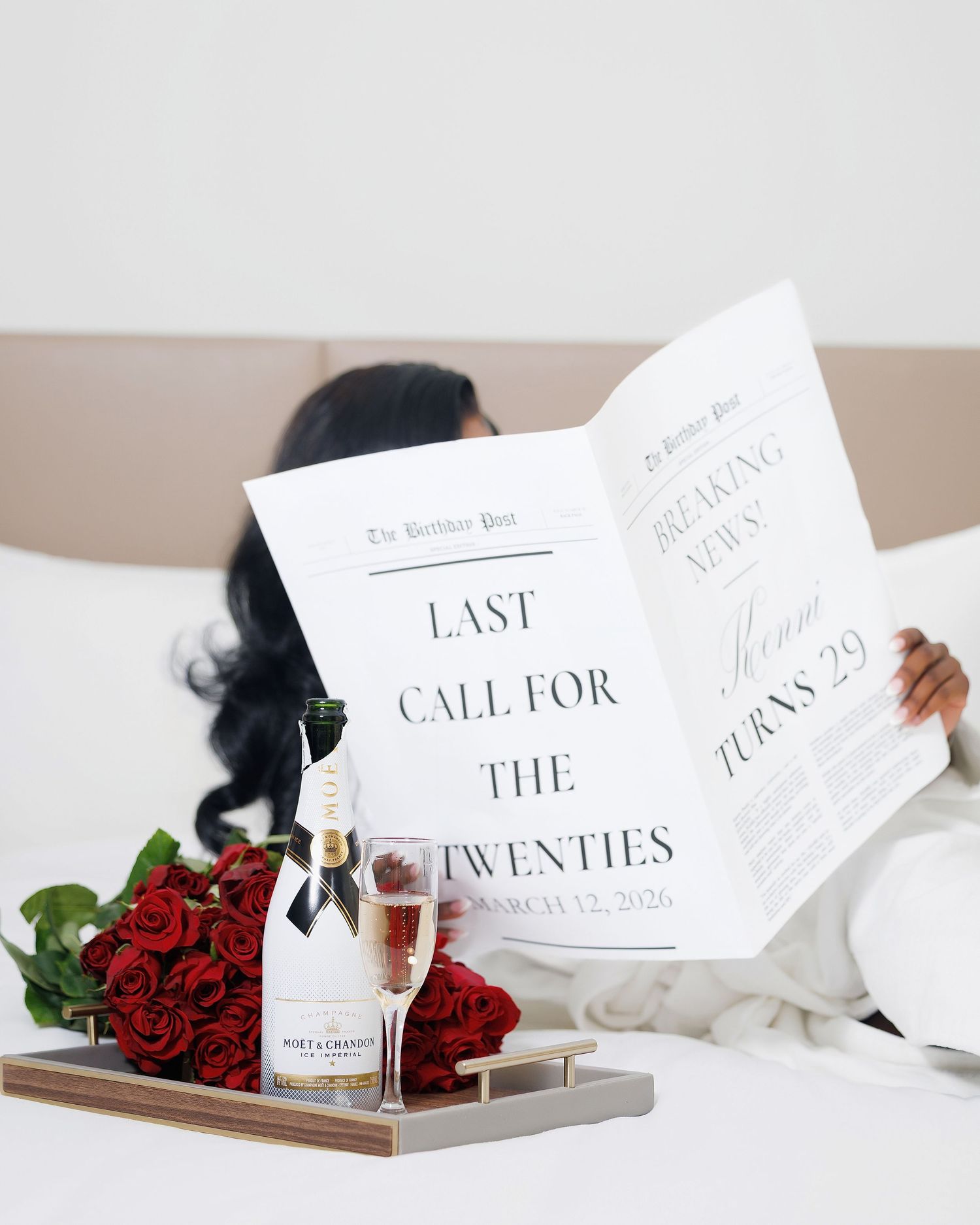 Woman in white robe reads birthday newspaper on bed with red roses and Moet champagne for 29th birthday celebration.