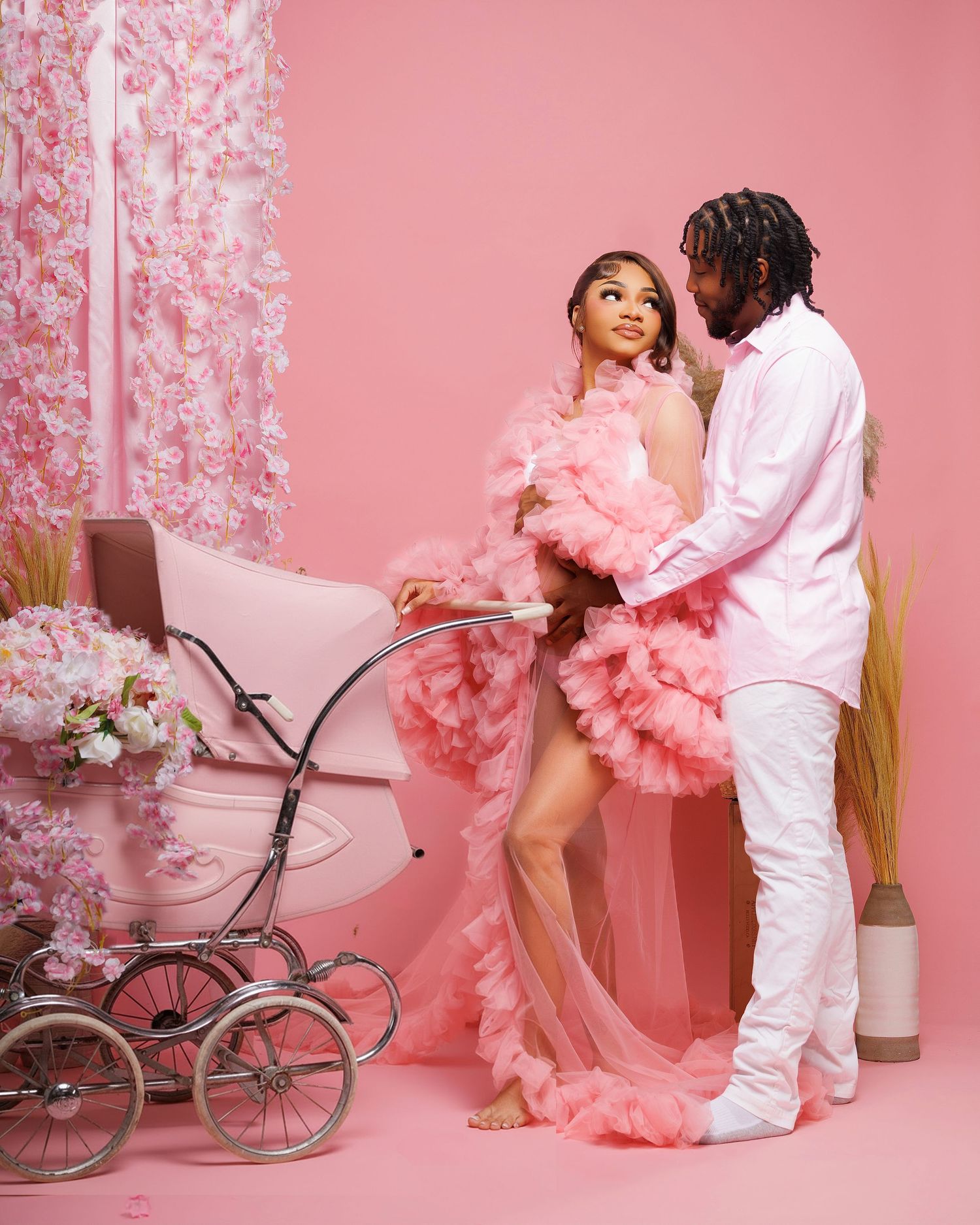 Couple in pink maternity photoshoot, woman in ruffled gown beside vintage pram, man in white outfit, pink floral backdrop.