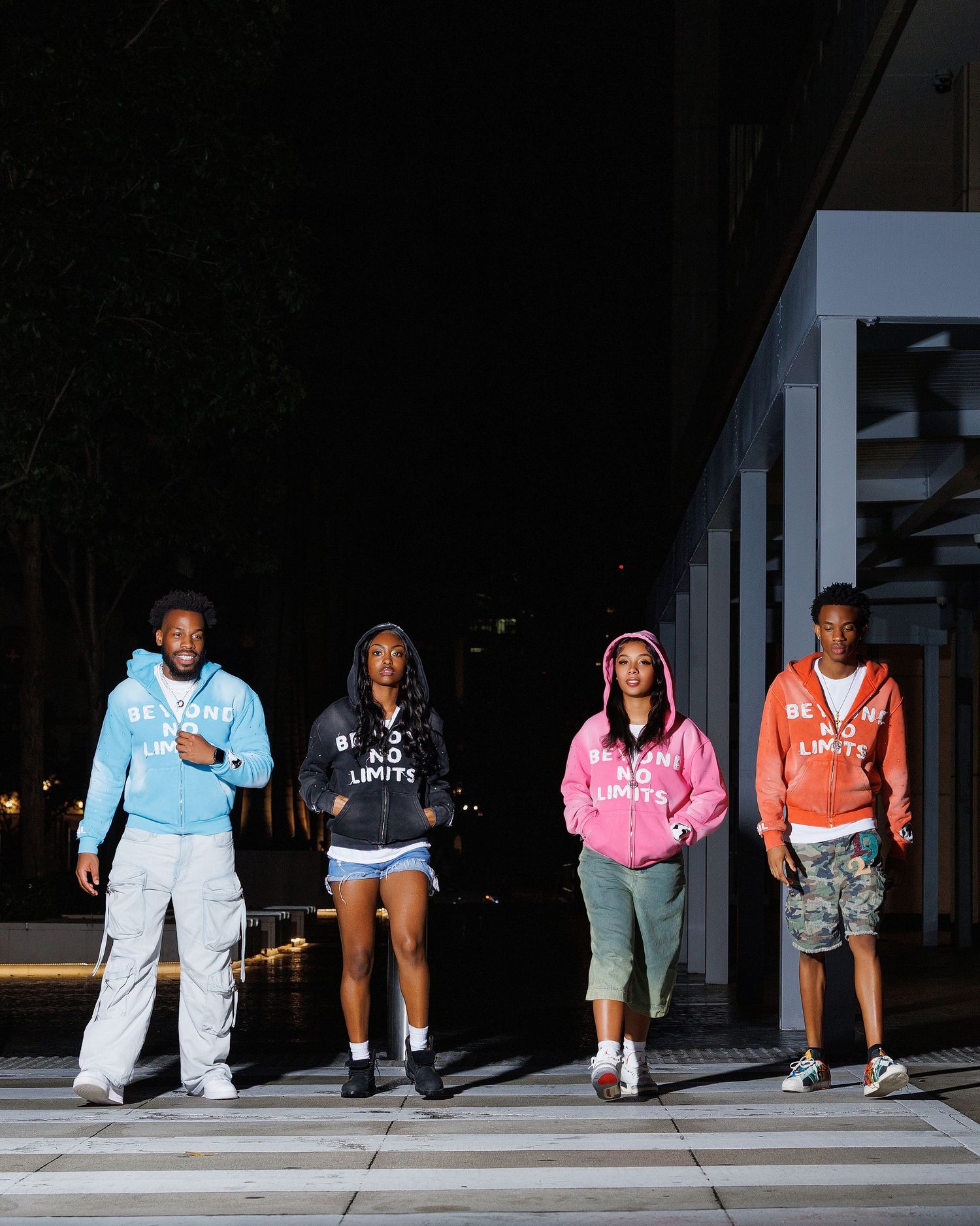 Four young people wearing matching Be No Limits hoodies in blue, black, pink, and coral pose outdoors at night.