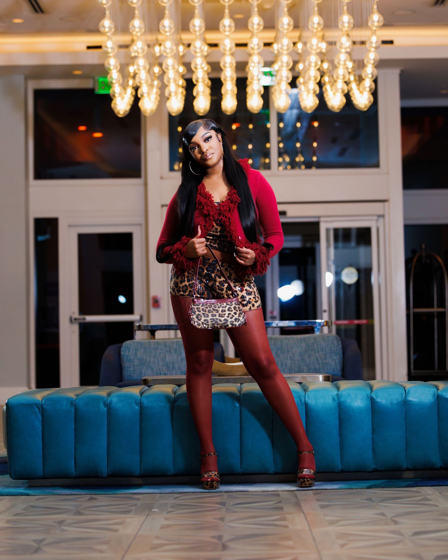 Woman in leopard print mini skirt and red jacket poses confidently in elegant hotel lobby under chandelier.