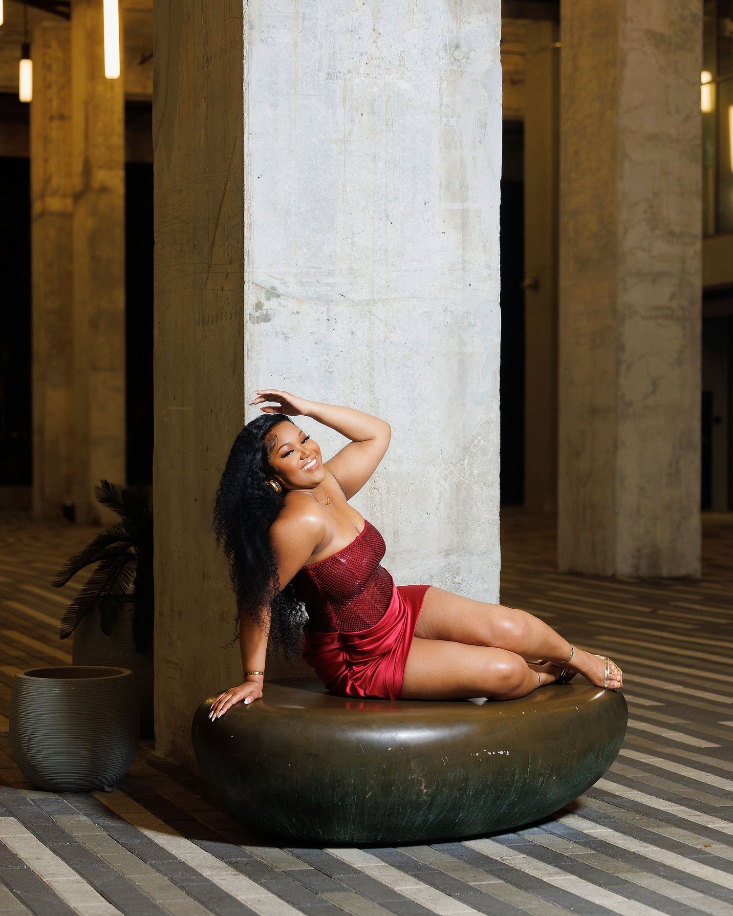 A woman in a red dress poses elegantly on a round ottoman beside a concrete column in a stylish interior.